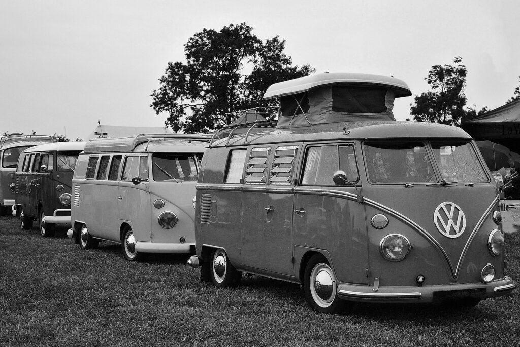 Eine Reihe alter Volkswagen Wohnmobile ist auf einer Wiese geparkt, mit Bäumen im Hintergrund. Das Schwarz-Weiß-Foto unterstreicht den nostalgischen Charme des Caravaning und fängt den Geist von früher und heute in diesen klassischen Fahrzeugen ein.