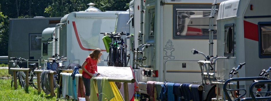 Eine Person in einem roten Hemd steht neben einer Reihe von geparkten Wohnmobilen und hängt Wäsche an einem Zaun auf einem der besten Caravaningplätze Deutschlands auf. Im Hintergrund sind mehrere Fahrräder angebracht und grünes Gras und Bäume zu sehen.
