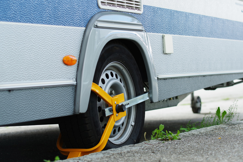 Eine Nahaufnahme eines Wohnmobils oder Anhängerrads, das mit einer gelben Radklemme gesichert ist, die zusätzlichen Einbruchschutz und Sicherheit bietet, geparkt neben einem Bordstein, um den herum etwas Gras wächst.