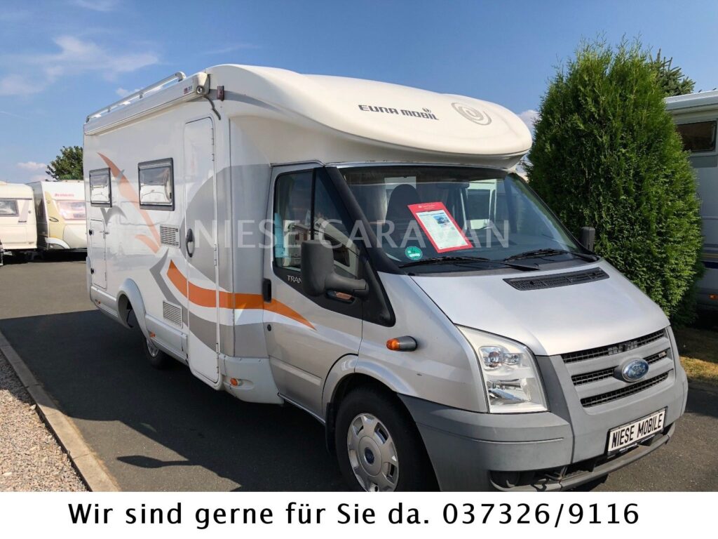 Ein silbernes Wohnmobil auf Ford-Basis mit orangefarbenen Akzenten steht auf einem gepflasterten Parkplatz, umgeben von anderen Wohnmobil-Marken. Auf dem vorderen Nummernschild ist ein Schild mit Kontaktinformationen und WIESE MOBIL angebracht.