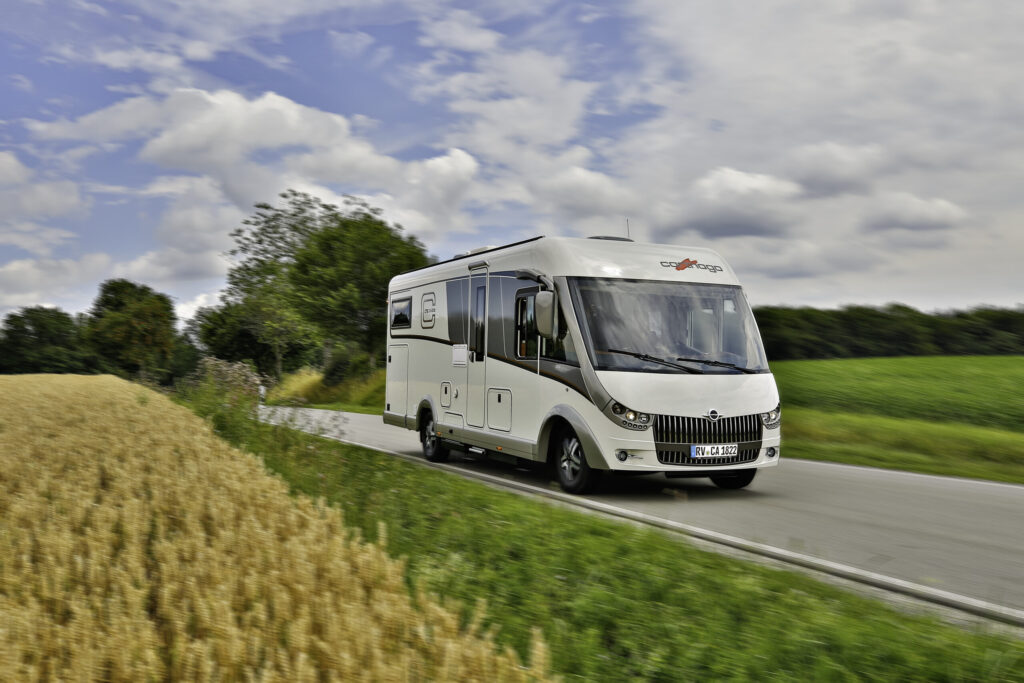 Ein weißes Wohnmobil fährt eine schmale Landstraße entlang, die von grünen Feldern und einem goldenen Weizenfeld unter einem teilweise bewölkten Himmel gesäumt wird. Im Hintergrund sind Bäume und entfernte Vegetation zu sehen.