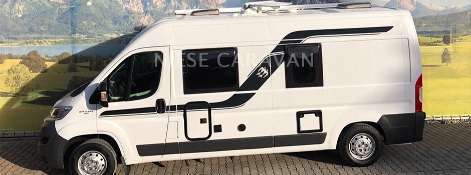 Ein weißer Kastenwagen mit schwarzer Grafik parkt auf einer gepflasterten Fläche vor einer malerischen Berg- und Wiesenkulisse. Dieses Wohnmobil verfügt über mehrere Fenster und Außenstaufächer für zusätzlichen Komfort.