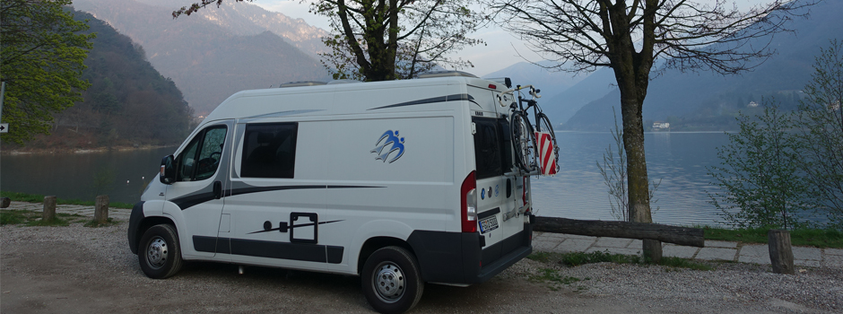 Ein weißes Wohnmobil auf einer Wohnmobil Reise parkt an einem ruhigen Seeufer, umgeben von Bäumen und Bergen, mit einem Fahrrad auf dem Rücken und ruhigem Wasser, in dem sich die heitere Landschaft der Reisetage 4-6 spiegelt.