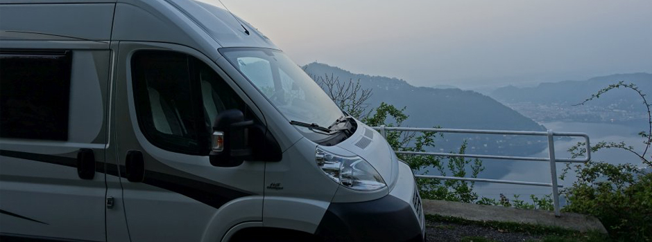 Ein weißes Wohnmobil steht auf einer Anhöhe neben einem Metallgeländer, mit Blick auf nebelverhangene Berge und einen See - genau das Richtige für alle, die auf einer Wohnmobil-Reise das Abenteuer unter einem wolkenverhangenen Himmel suchen.