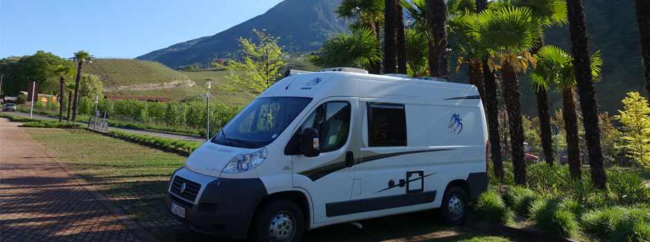 Ein weißes Wohnmobil parkt auf einer Ziegelsteinstraße neben hohen Palmen, mit grünen Hügeln und einem Berg im Hintergrund unter einem klaren blauen Himmel - eine ideale Szene für jede Wohnmobil Reise oder einen Eintrag in Ihr Reisetagebuch.