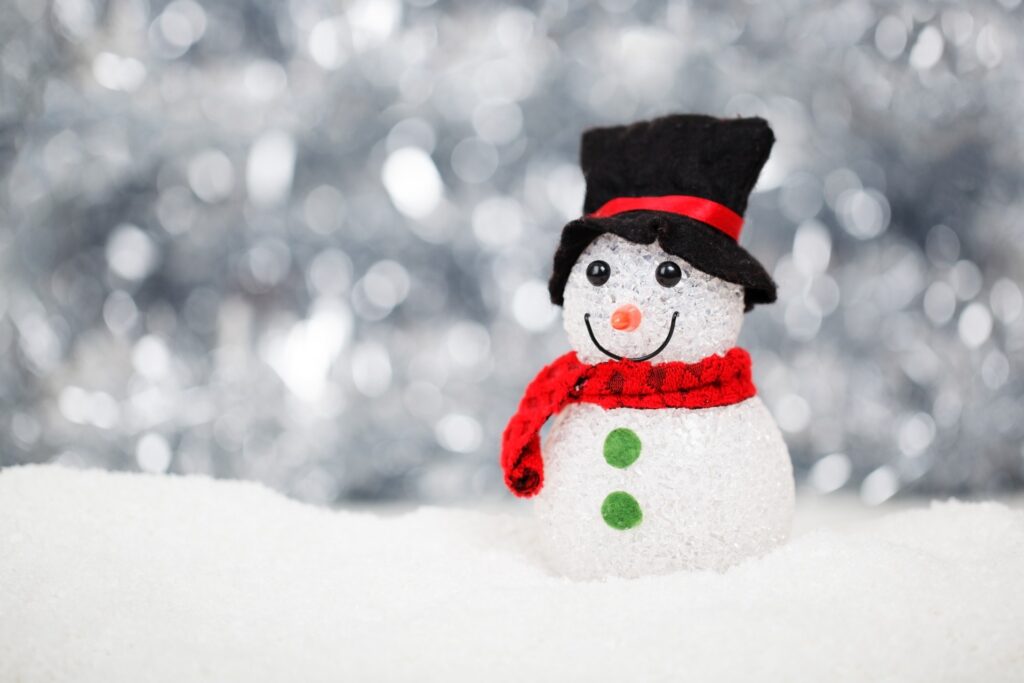 Eine kleine Schneemann-Figur mit schwarzem Zylinder, rotem Schal, grünen Knöpfen und einem lächelnden Gesicht steht auf weißem Schnee mit verschwommenem Silberhintergrund - perfekt, um Weihnachten im Wohnmobil festliches Flair zu verleihen.