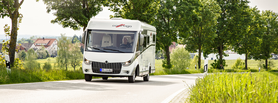 Ein weißes Carthago Reisemobil fährt auf einer kurvenreichen Landstraße, die von grünem Gras und Bäumen gesäumt ist. Im Hintergrund sind an einem sonnigen Tag Häuser und Felder zu sehen. Entdecken Sie Top Wohnmobil Marken für Ihr nächstes Abenteuer.