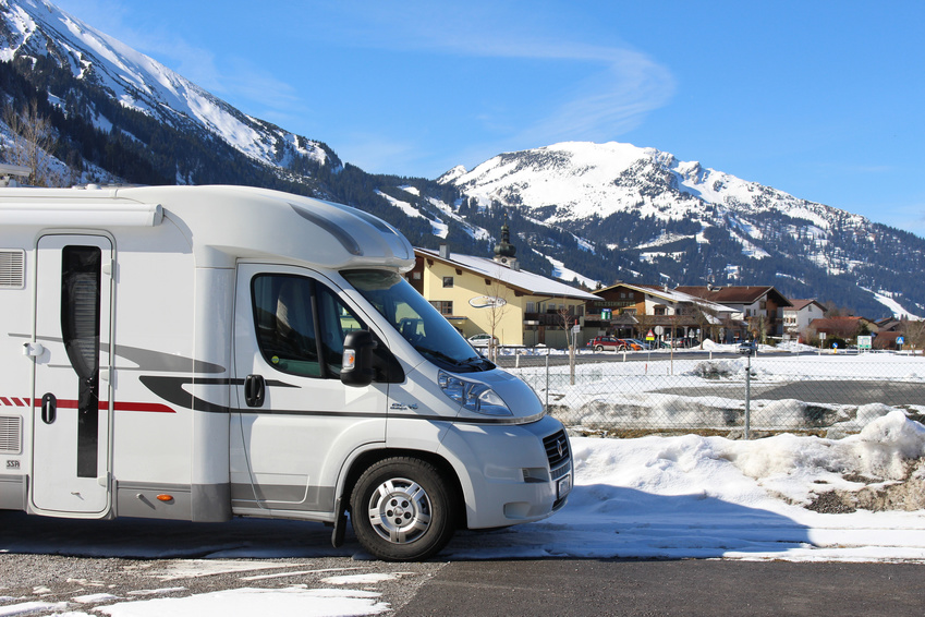 Ein weißes Wohnmobil parkt auf einer verschneiten Straße mit Bergen und Häusern im Hintergrund unter einem klaren blauen Himmel, bereit für Winterabenteuer mit extra Pflege.