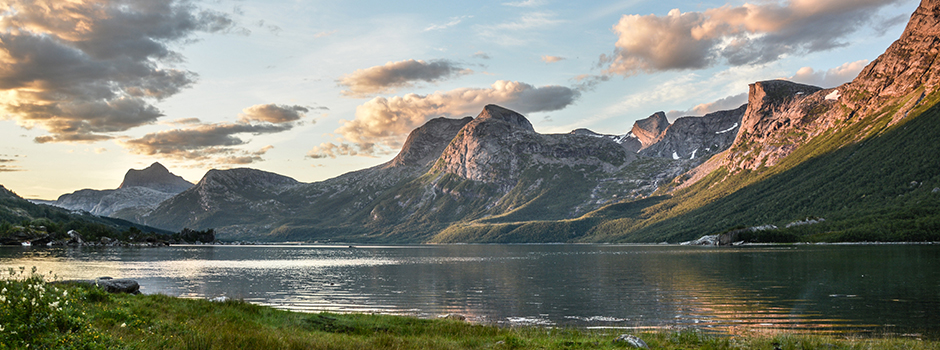 Eine Gebirgskette mit scharfen Gipfeln und grünen Hängen spiegelt sich in einem ruhigen See unter einem teilweise bewölkten Himmel - ideal für Wohnmobil Touren in Norwegen. Der grasbewachsene Vordergrund und die bewaldeten Flächen säumen das Wasser, alles sanft beleuchtet vom Sonnenlicht.
