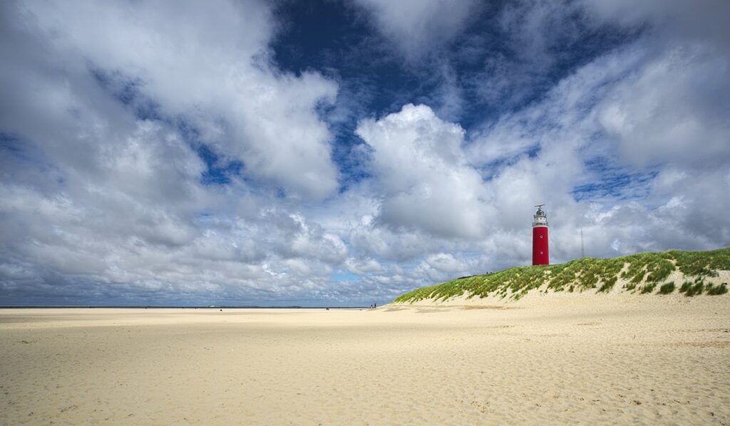 Ein rot-weißer Leuchtturm steht auf einer grasbewachsenen Sanddüne unter einem wolkenblauen Himmel und überblickt einen breiten, leeren Sandstrand - perfekt für einen Urlaub an der Nordsee mit traumhaften Wohnmobilstellplätzen in unmittelbarer Nähe.