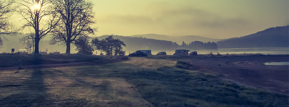 Frühmorgendliche Landschaft in Mitteldeutschland: Nebelschwaden ziehen über Bäume und mehrere Autos, die an einem See geparkt sind. Die Sonne wirft lange Schatten auf die Wiese am Wasser, perfekt für Camping am See, mit den Hügeln im Hintergrund.