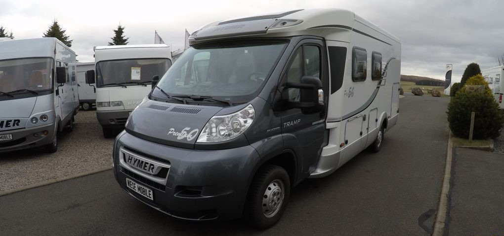 Ein grau-weißer Hymer Tramp, eines der beliebten teilintegrierten Wohnmobile, steht auf einem gepflasterten Parkplatz zwischen anderen Wohnmobilen unter einem bewölkten Himmel, umgeben von Bäumen und Büschen.