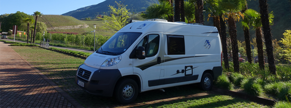 Ein weißes Wohnmobil, geparkt an einem gemauerten Weg, umgeben von Palmen und Grün, mit Bergen und Weinbergen im Hintergrund unter einem klaren blauen Himmel - perfekt für eine unvergessliche Reise.