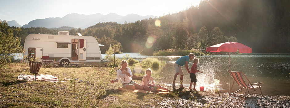 Eine vierköpfige Familie genießt ein Picknick am See in der Nähe eines weißen Dethleffs-Wohnmobils; zwei Erwachsene sitzen auf einer Decke, während zwei Kinder am Ufer mit Liegestühlen und einem roten Sonnenschirm spielen, mit Bergen und Bäumen im Hintergrund.